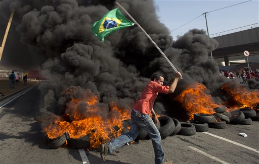 A member of the Homeless Workers Movement carries a Brazilian flag past burning tires during a protest against the money spent on the World Cup near Itaquerao stadium which will host the international soccer tournament's first match in Sao Paulo, Brazil, Thursday, May 15, 2014. Brazilians are angry at the billions spent to host the World Cup, much of it on 12 ornate football stadiums, one-third of which critics say will see little use after the big event. (AP Photo/Andre Penner)