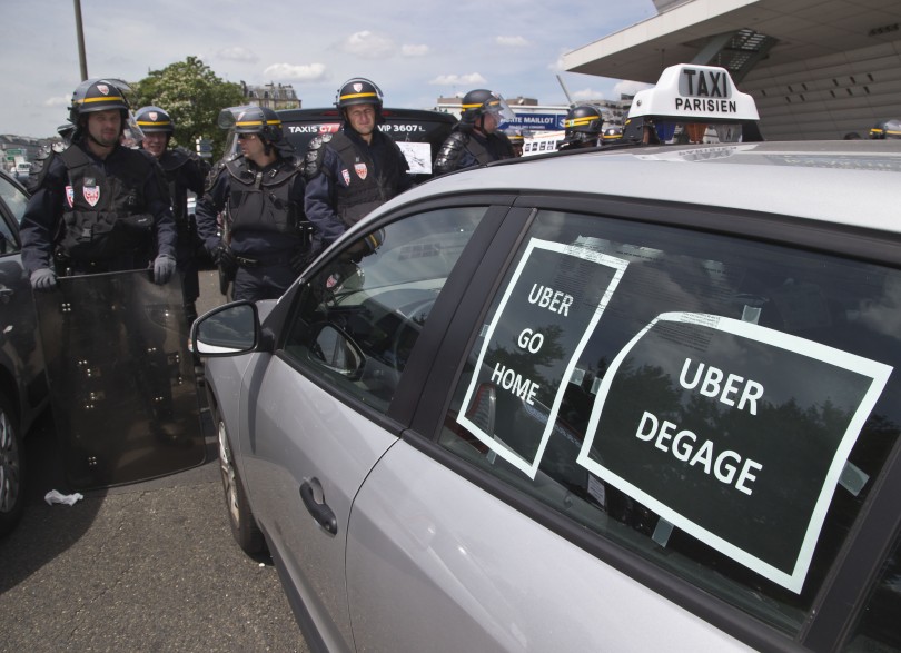 Riot police officers stand next to a cab with posters which reads, "Uber go home" and "Uber get out out," during a taxi drivers demonstration in Paris, France, Thursday, June 25, 2015. (Michel Euler/AP)