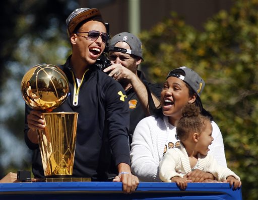 Golden State Warriors guard Stephen Curry, left, holds the Larry O'Brien Championship Trophy as he rides a bus with his daughter Riley, front right, and wife Ayesha as the procession turns onto Broadway in downtown Oakland, Calif., during a parade for the team's winning of the NBA basketball championship Friday, June 19, 2015. (Karl Mondon/San Jose Mercury News via AP)