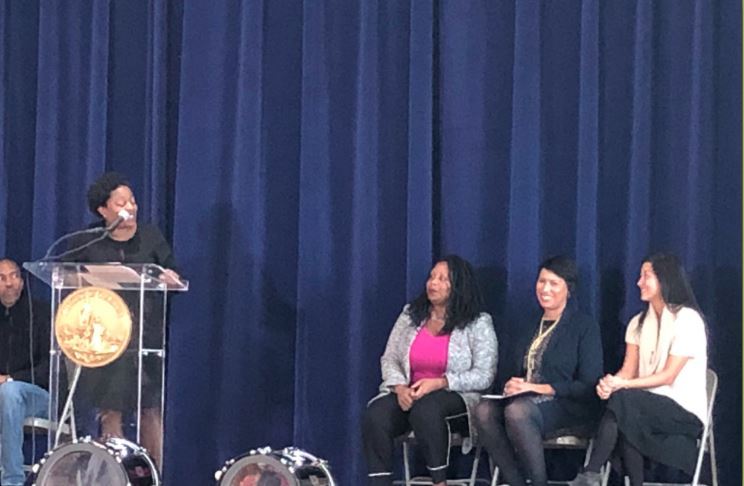 DCPS Interim Chancellor Dr. Amanda Alexander speaks at an assembly presenting Principal Maisha Riddlesprigger (seated far left) of Ketcham Elementary with the 2019 DCPS Principal of the Year Award. Riddlesprigger is seated next to D.C. Mayor Muriel Bowser and District of Columbia State Superintendent Hanseul Kang. 
