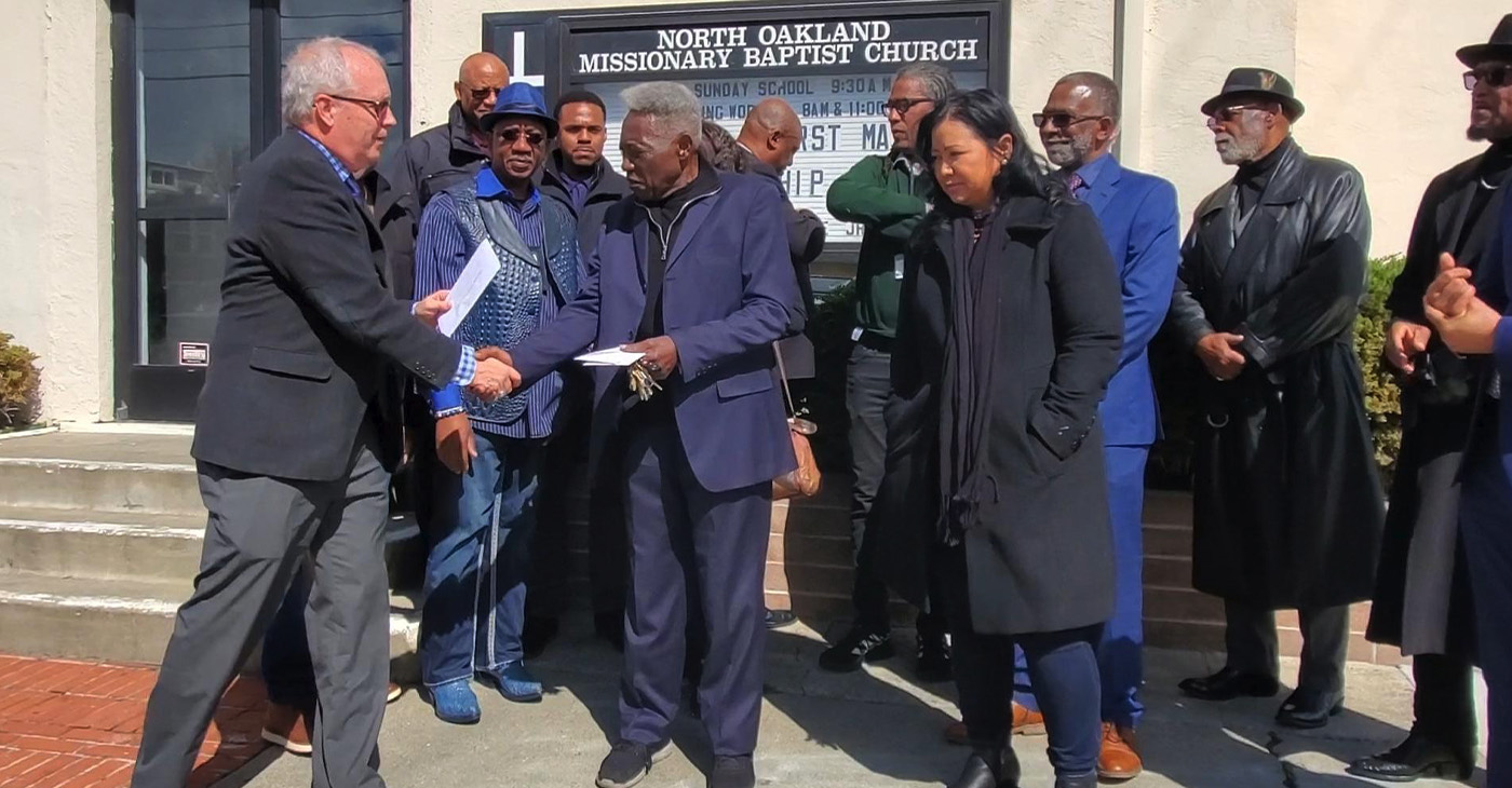 As fellow pastors look on, Tim Hopkins of Lakeshore Avenue Baptist Church presents a $1,000 check to Pastor Sylvester Rutledge of North Oakland Missionary Baptist Church (NOMBC) outside the church in Oakland, California. Photo By Carla Thomas