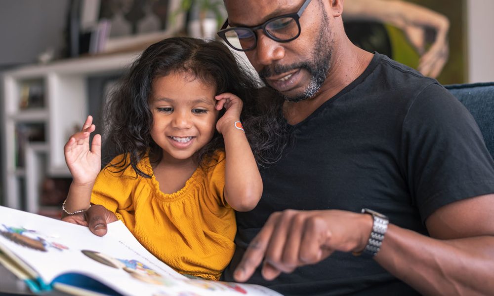A loving father sits on the couch at home and reads a storybook to his preschool age daughter.