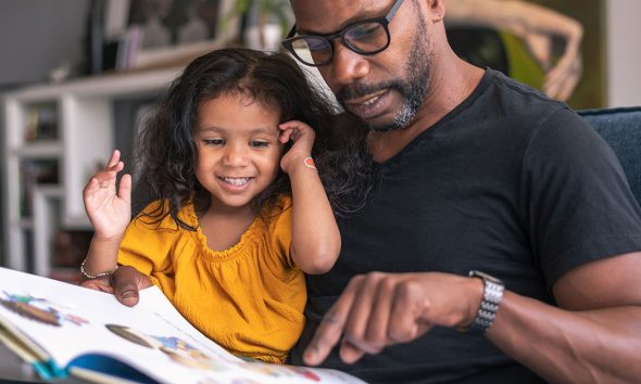 A loving father sits on the couch at home and reads a storybook to his preschool age daughter.