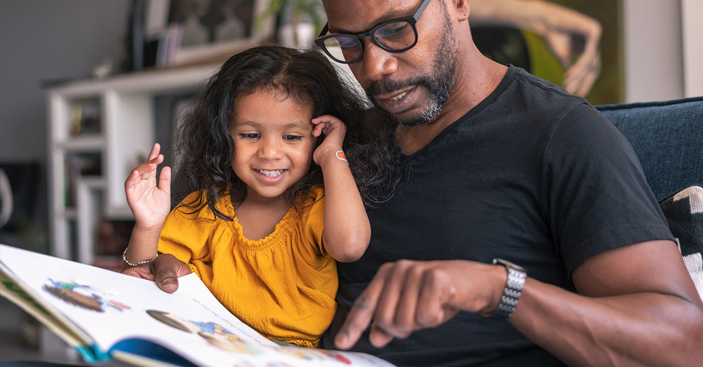 A loving father sits on the couch at home and reads a storybook to his preschool age daughter.
