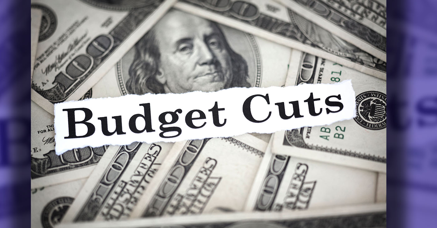 The words "Budget Cuts" with hundred dollar bills in the background.