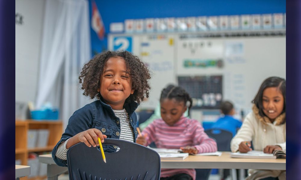 A super cute African American elementary school girl smiles at the camera as she sits at her desk.