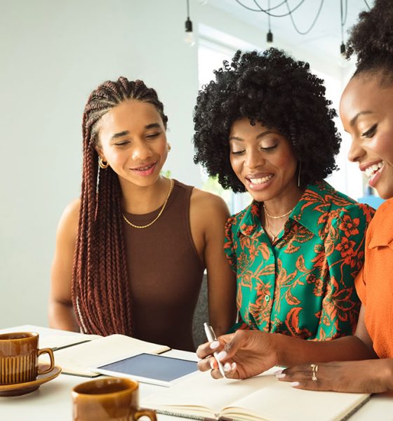 Teamwork concept. Three beautiful black women wearing dresses sitting at the table in an office and discussing over project.