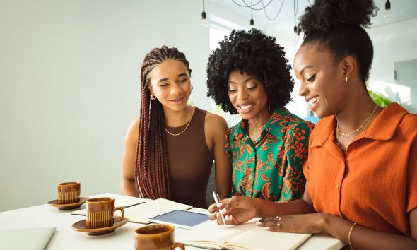 Teamwork concept. Three beautiful black women wearing dresses sitting at the table in an office and discussing over project.