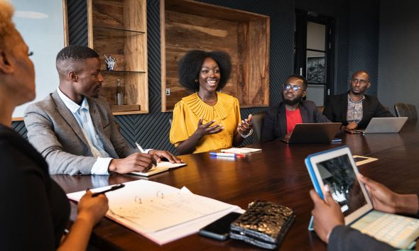 View between colleagues with focus on African professional in early 30s looking across conference table at young associate, smiling and gesturing as she speaks. (Photo by Johnny Greig)