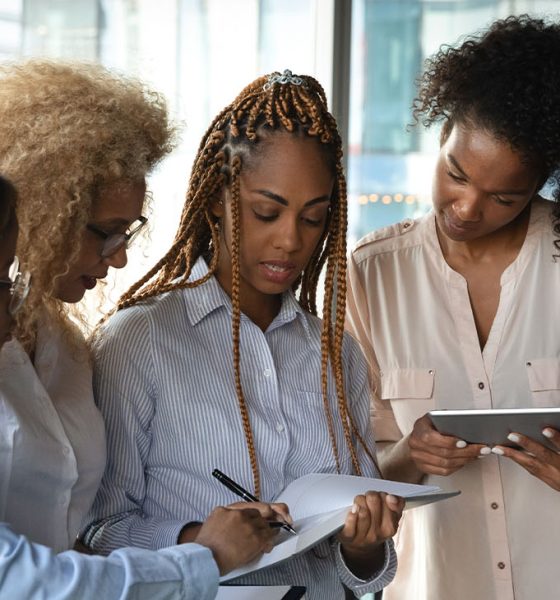 Informal brainstorming. Supportive mixed race females workers consult colleague help to find mistake in report. Experienced women managers share skills with intern during work break at office corridor