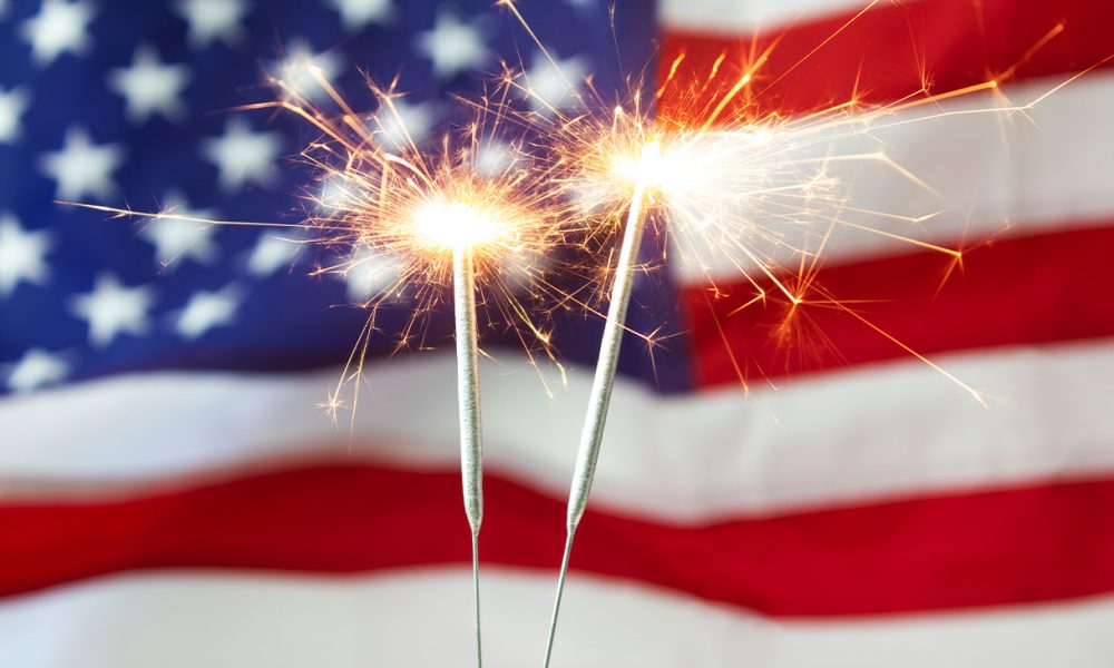 Two bright burning sparklers with an American flag in the background. 4th of July celebrations.