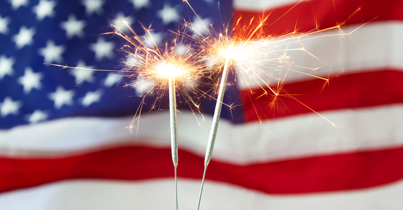 Two bright burning sparklers with an American flag in the background. 4th of July celebrations.