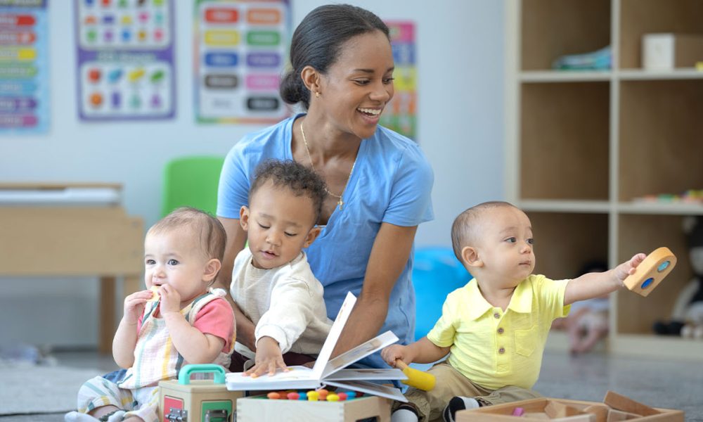 A daycare teacher sits on the floor of the classroom with one child on her lap and one on either side of her, as they play together with toys. The children are each dressed casually and engaged with different activities.