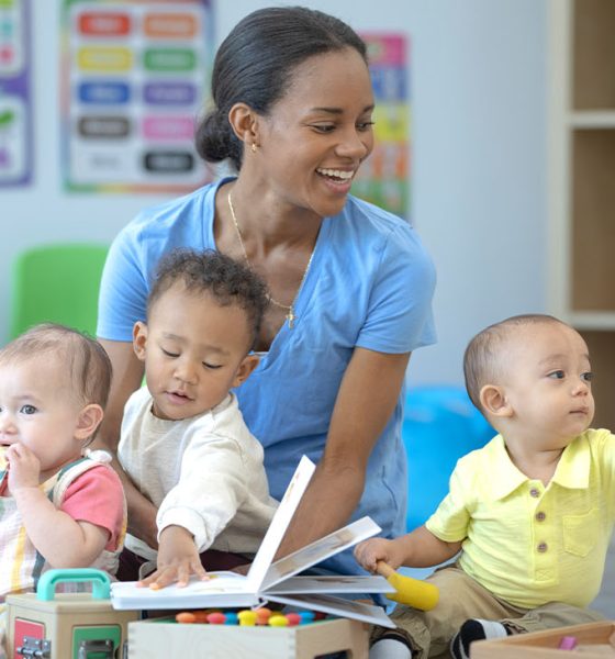 A daycare teacher sits on the floor of the classroom with one child on her lap and one on either side of her, as they play together with toys. The children are each dressed casually and engaged with different activities.