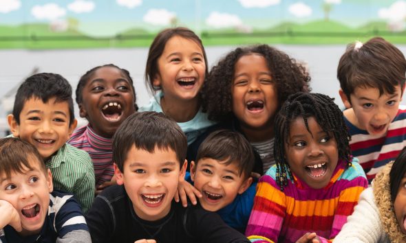 A group of diverse kids smile in this portrait. They are stacked on top of each other while cuddling in close and showing how happy they are.