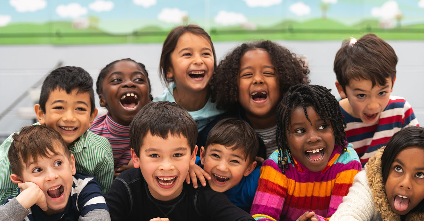 A group of diverse kids smile in this portrait. They are stacked on top of each other while cuddling in close and showing how happy they are.