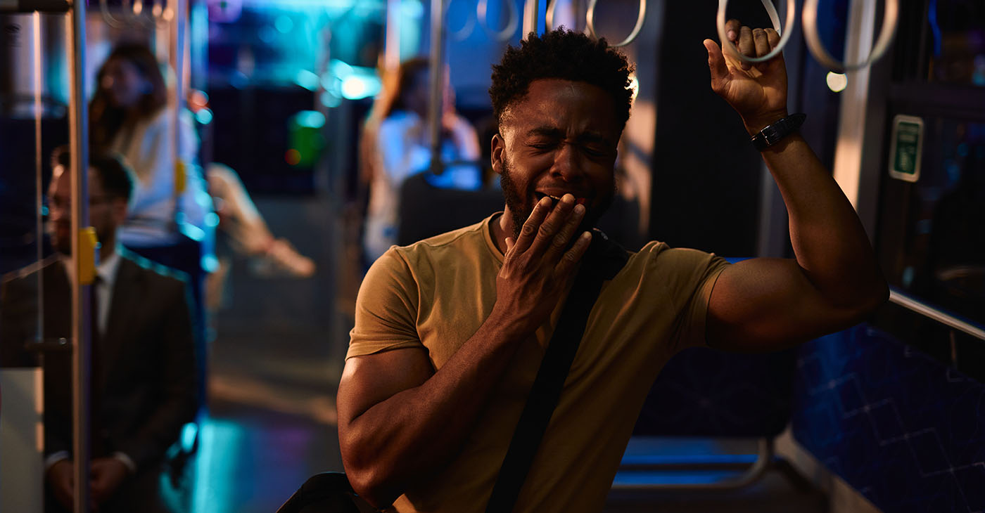 Sleepy young man covers his mouth while yawning and holding onto the railing of a night bus, other passengers are visible in the background (Photo by Igor Suka)