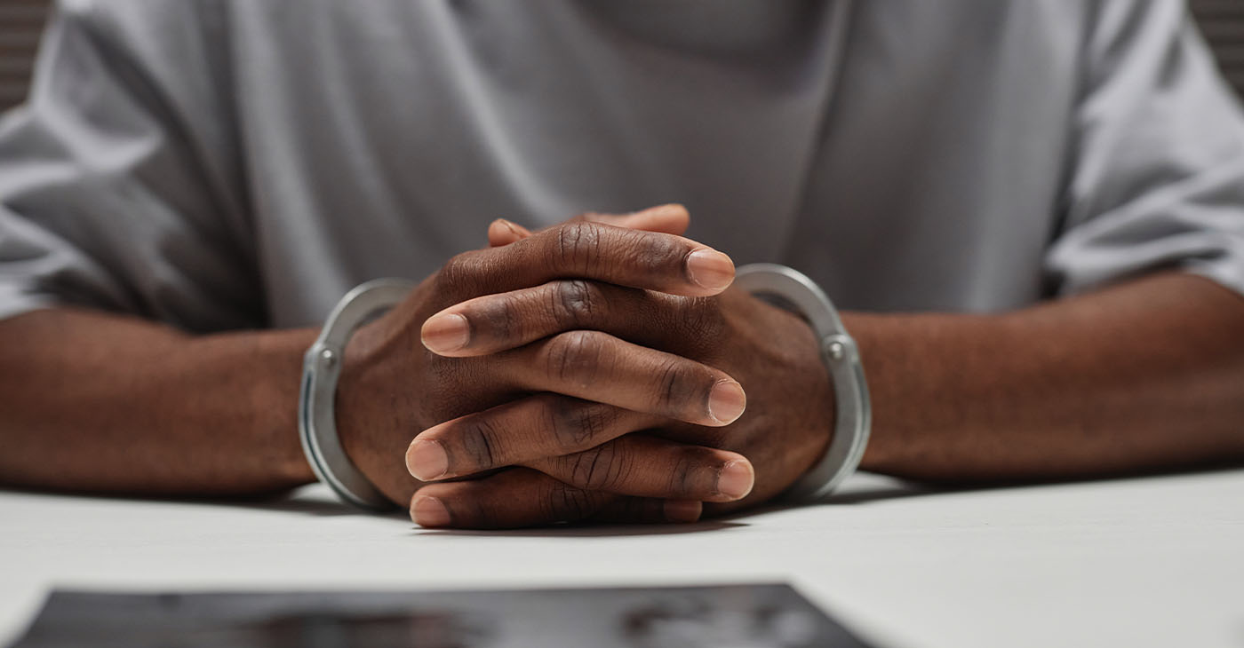 Close up of anonymous black man wearing handcuffs sitting at table in police department with hands clasped, copy space