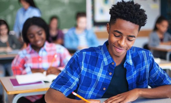Smiling male student writing in classroom. Teenage boy is concentrating at high school. He is sitting at desk.