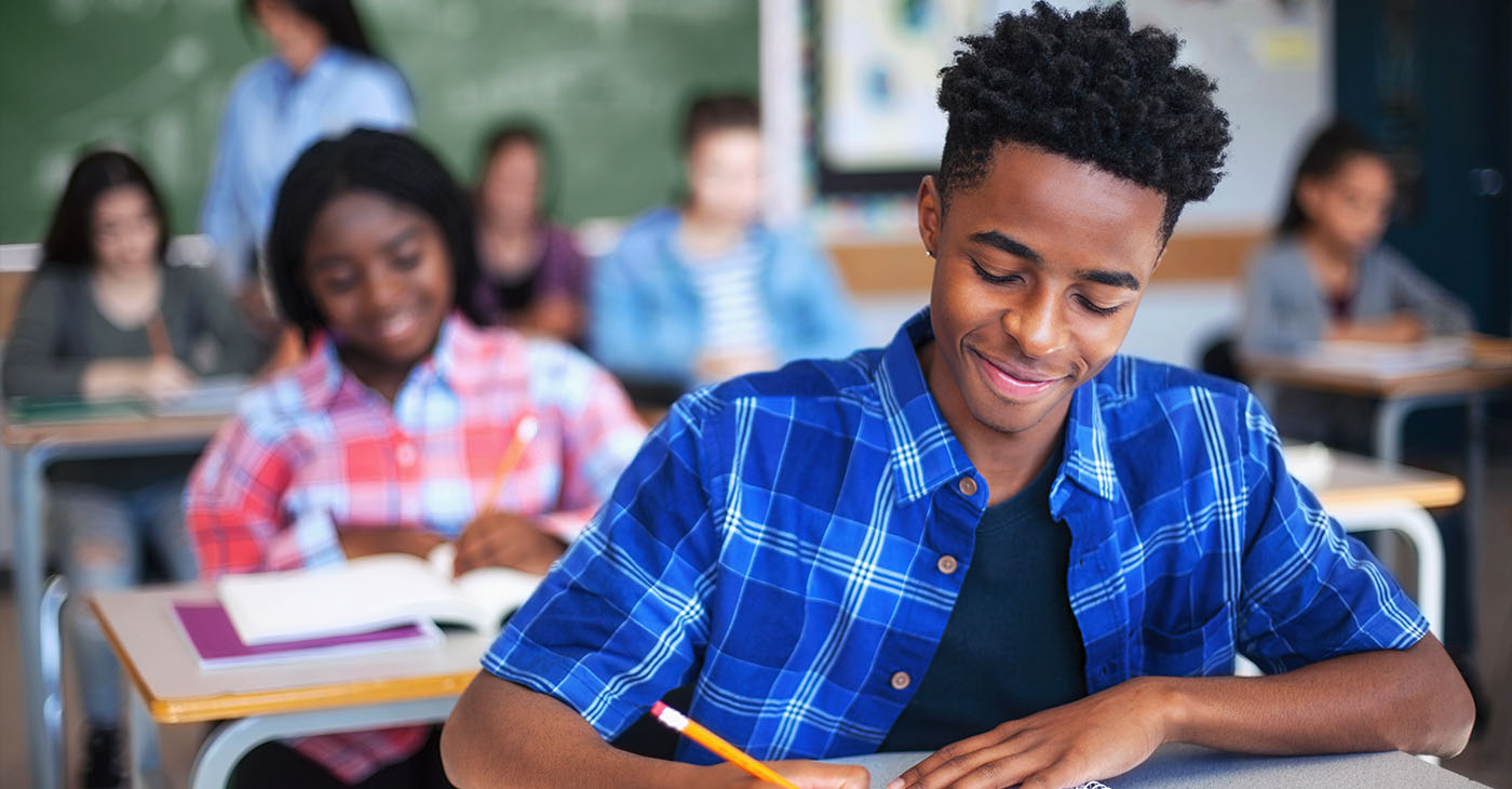 Smiling male student writing in classroom. Teenage boy is concentrating at high school. He is sitting at desk.