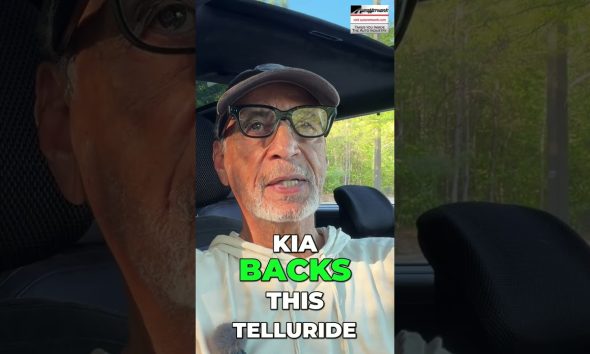 Man wearing cap and glasses sits in a car, outdoor light behind him; overlaid text reads 'KIA BACKS THIS TELLURIDE'.