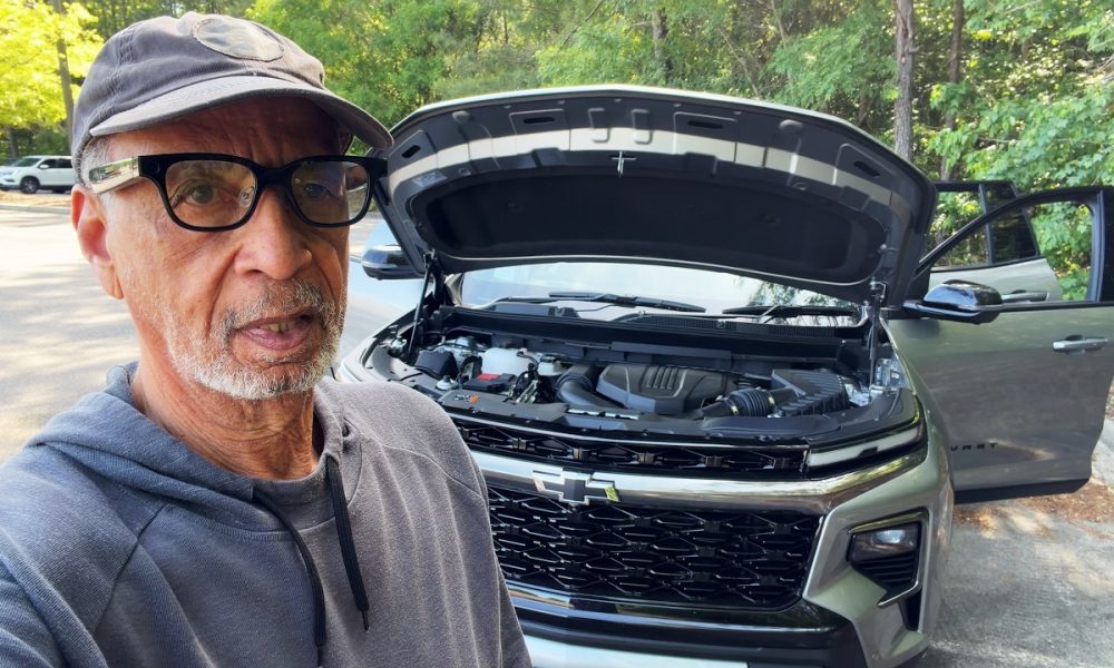 Older man wearing glasses and a cap takes a selfie beside a gray SUV with the hood up in a parking lot.