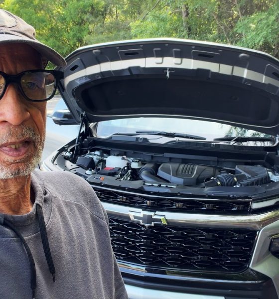 Older man wearing glasses and a cap takes a selfie beside a gray SUV with the hood up in a parking lot.