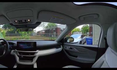 Interior of a modern car viewed from the passenger side, showing dashboard with touchscreen, steering wheel, and sunroof, with street outside the windows.