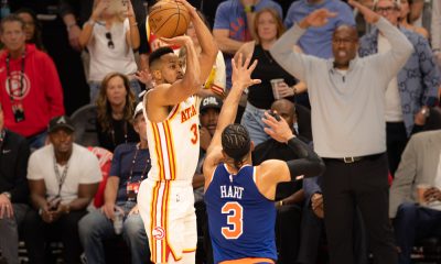 Basketball player in a white Atlanta Hawks uniform (No. 3) shoots over a defender in blue (No. 3) with a crowded arena background nearby.
