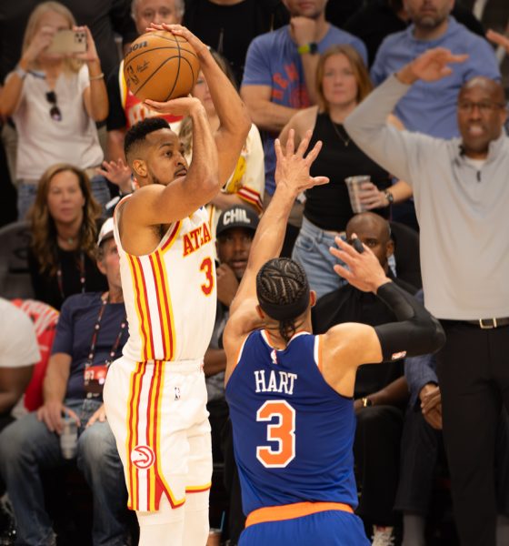 Basketball player in a white Atlanta Hawks uniform (No. 3) shoots over a defender in blue (No. 3) with a crowded arena background nearby.