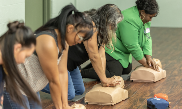 People kneeling on a gym floor practicing chest compressions on CPR manikins during a hands-on training session, with a green-clad instructor nearby.